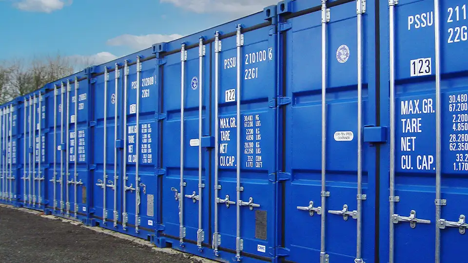 Row of bright blue shipping containers at a U Can Store It self storage site, each labeled with unit numbers and capacity details, set on a gravel surface under a blue sky.