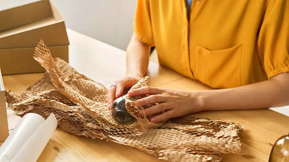 Person wrapping a glass bottle in brown honeycomb paper for protective packaging, with cardboard boxes on a wooden table, preparing items for self storage.